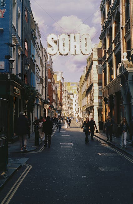 A lively street scene in Soho, London, with a large illuminated 'SOHO' sign hanging above the pedestrianized street. The street is lined with multi-story buildings featuring a mixture of traditional a