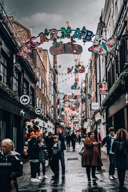A lively street scene in Soho, London, with a large illuminated 'SOHO' sign hanging above the pedestrianized street. The street is lined with multi-story buildings featuring a mixture of traditional a